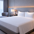 A neatly made bed with crisp white linens and two white pillows in a modern hotel-style bedroom with a gray upholstered headboard, bedside lamp, chair, and window.